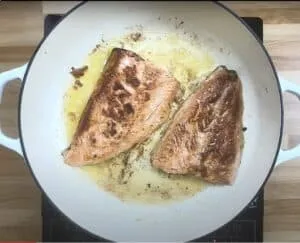 Two seared salmon fillets in a white pan with butter.